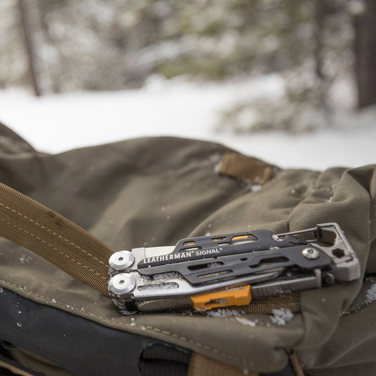 A gray Leatherman Signal closed on a backpack surrounded by snow.
