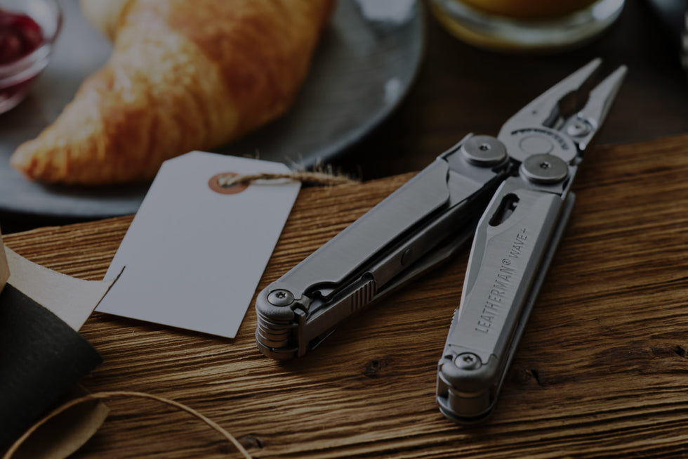 A pair of Leatherman Wave rests beside a croissant on a wooden table, showcasing an unusual juxtaposition of tools and food.